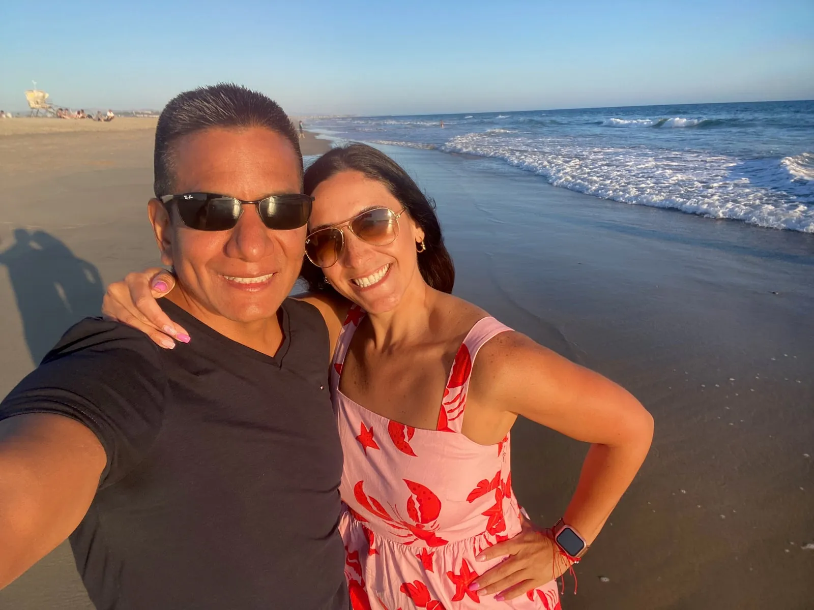 Katherine and Isaac smiling in sunglasses on the beach, the ocean behind them