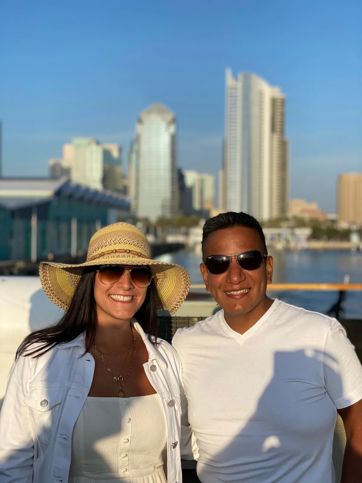Katherine and Isaac in sunglasses by the water, a city skyline behind them