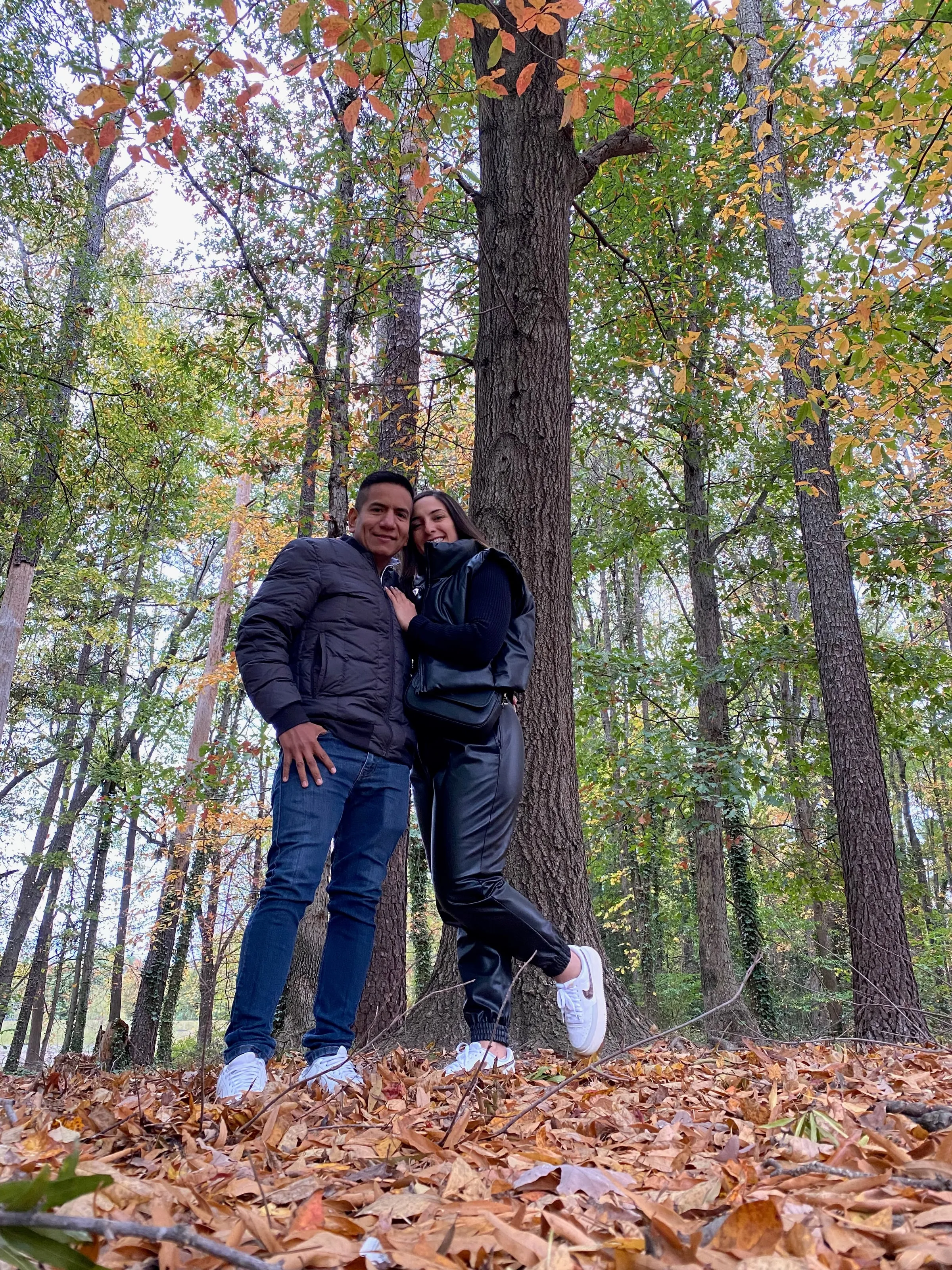 Katherine and Isaac embracing beneath tall autumn trees, the ground covered in fallen leaves